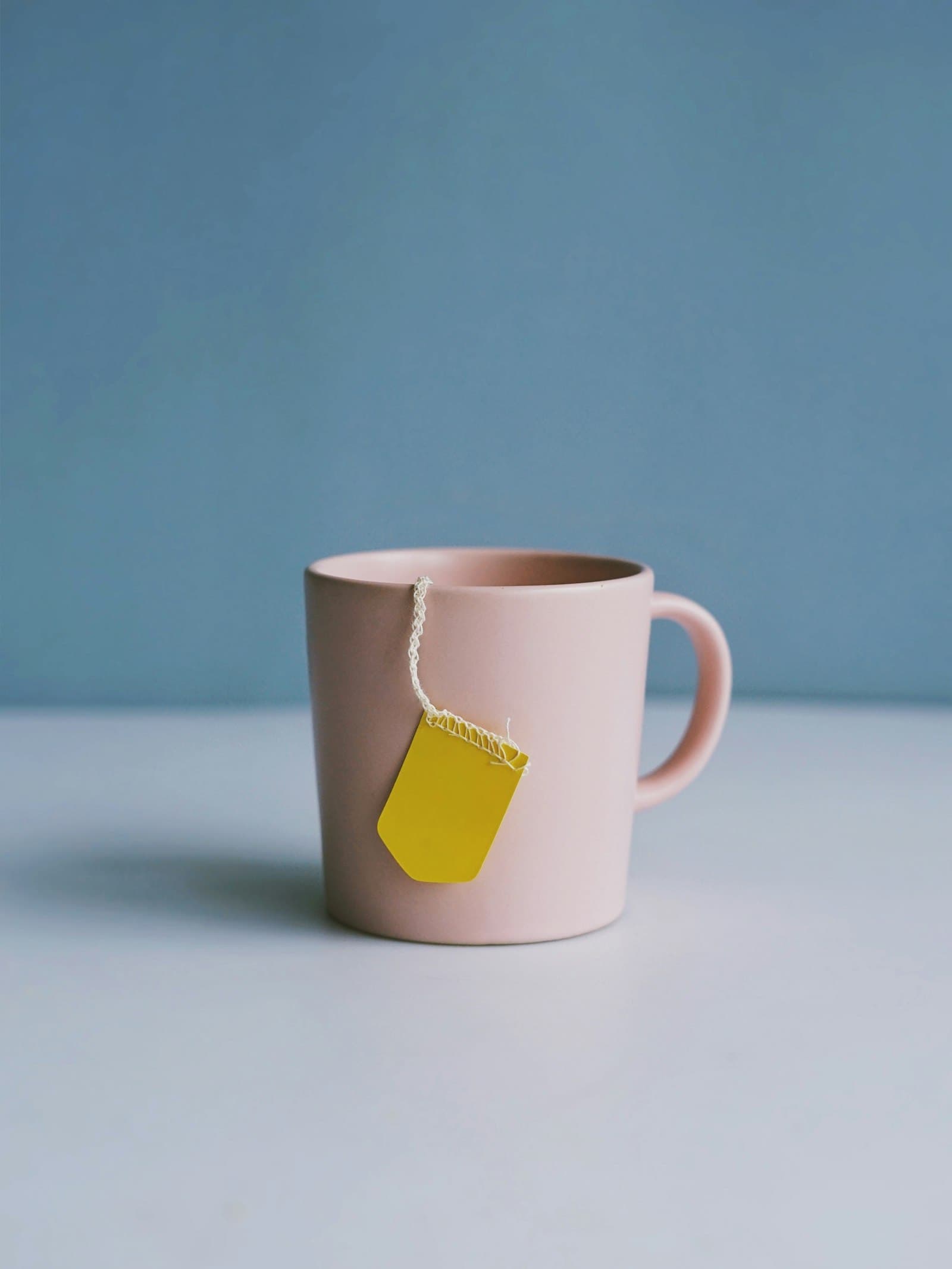 A pale blush ceramic mug set against a dusty blue backdrop, a yellow tea-bag tag hanging over the rim by its string, soft indirect light.