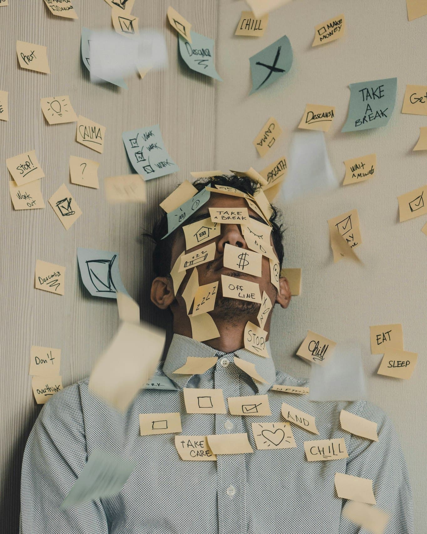 A person in a pale blue shirt against a wood-panelled wall, their face obscured by dozens of yellow and pale blue sticky notes bearing handwritten reminders like 'take a break', 'stop', 'chill' and 'don't overthink'.