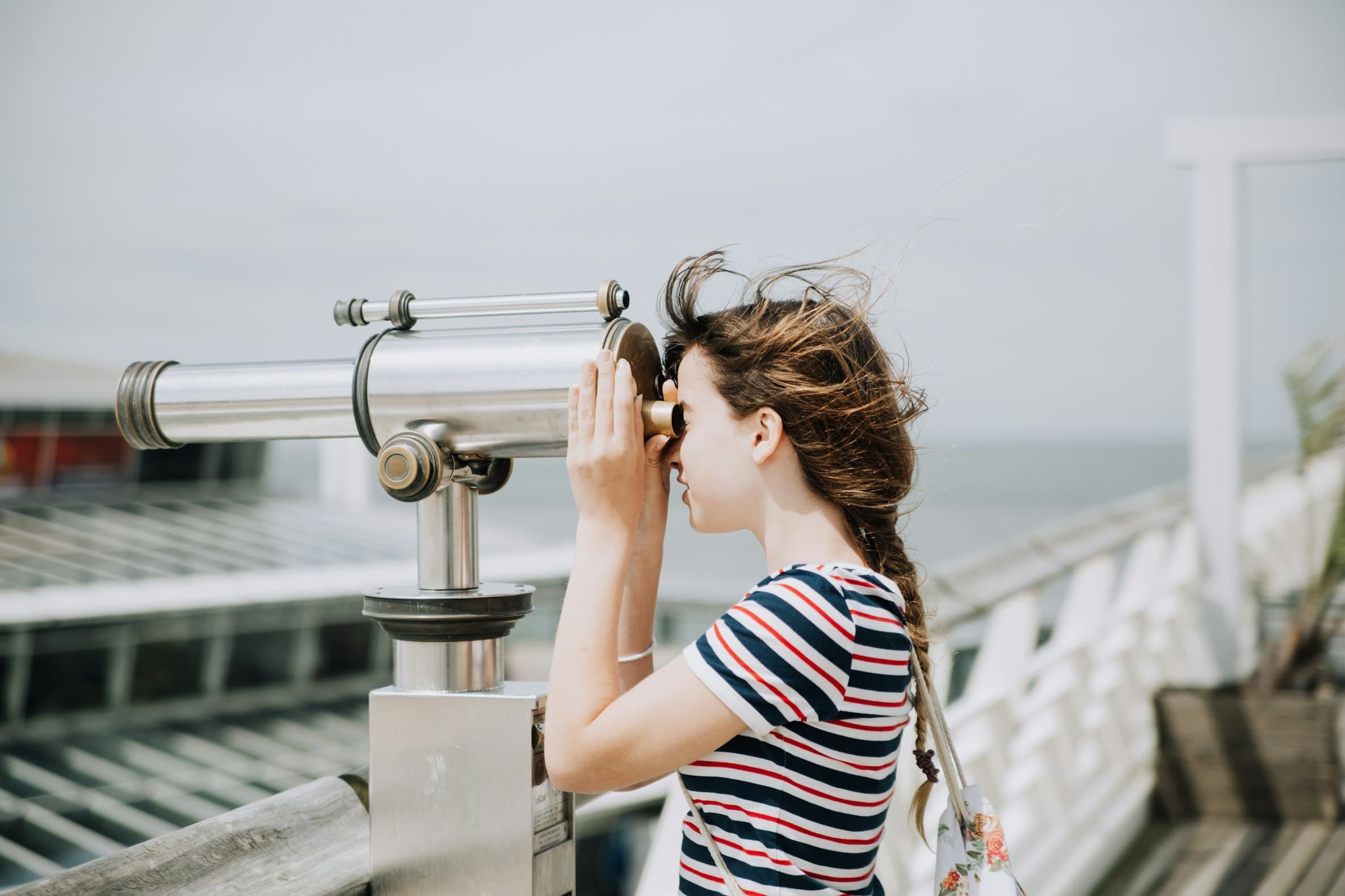 A child in a striped t-shirt leaning into a coin-operated seaside telescope, looking carefully through the eyepiece.