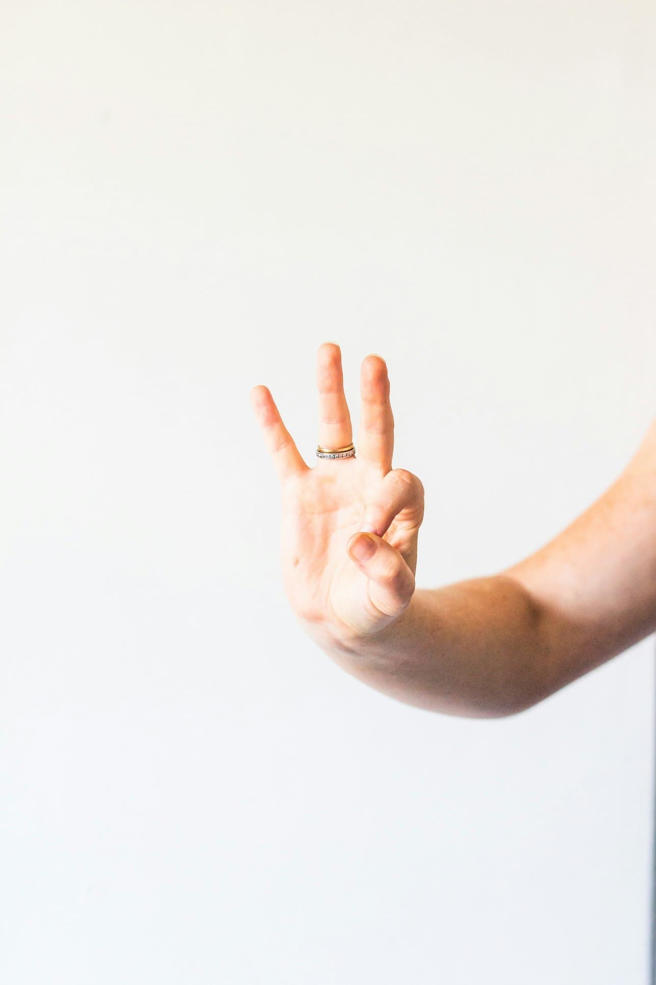 A hand raised against a soft white backdrop with three fingers held up, a slim gold ring at the base of two of them.