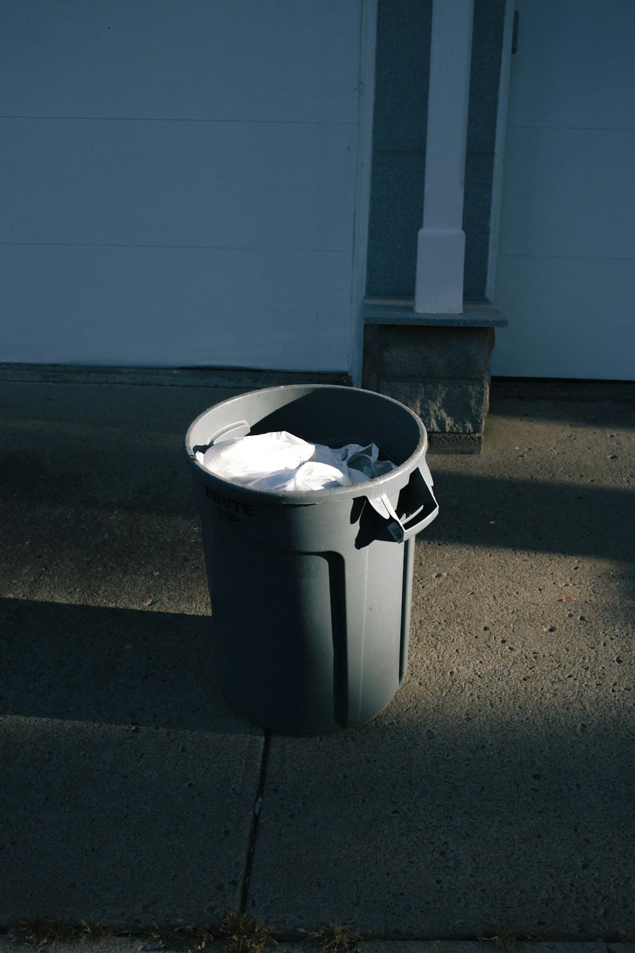 A large grey plastic bin on a sunlit pavement, a white bin bag spilling over the rim, a sharp shadow falling across the pale blue wall behind.