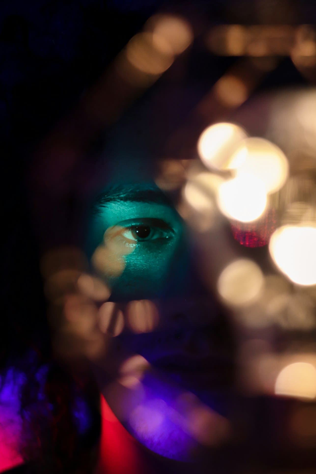 A single eye lit in teal by a handheld light, the rest of the face in shadow, warm bokeh lights blurring across the right half of the frame.