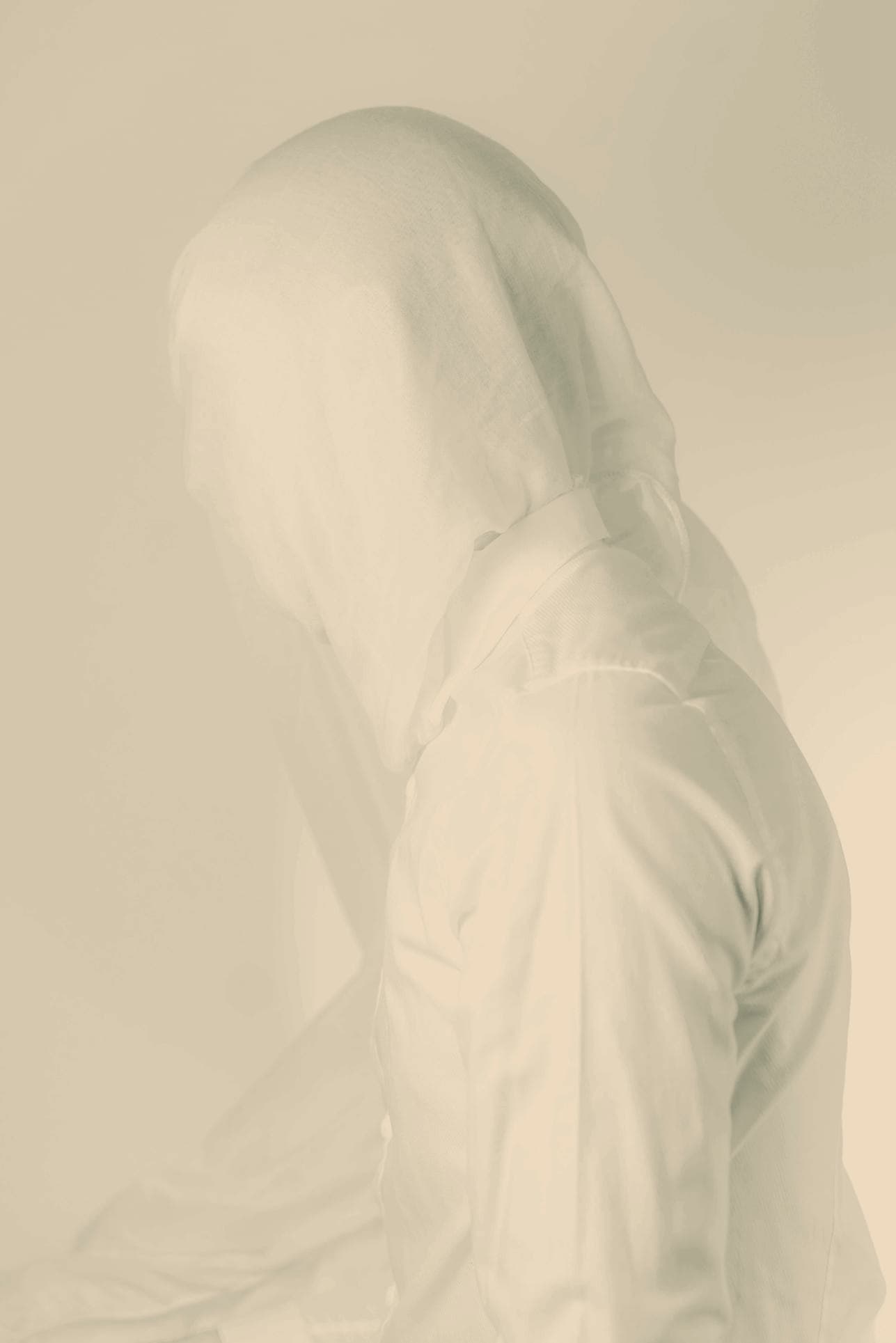 A figure in a white shirt with a hood pulled over their head, face obscured, photographed against a pale wall in soft daylight.