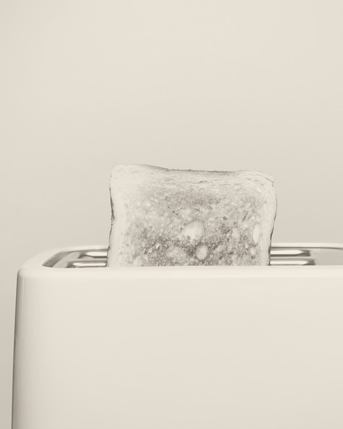 A single slice of toast sitting proud of a white two-slice toaster against a pale grey background.