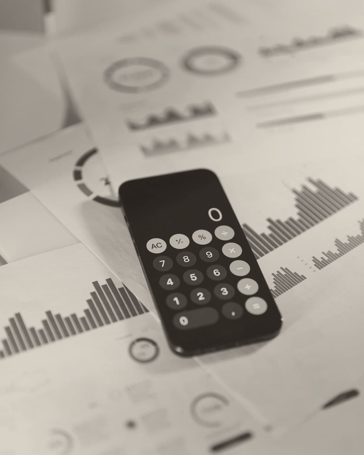 A phone showing the calculator app laid across printed reports with coloured bar charts and pie charts, daylight falling softly from above.