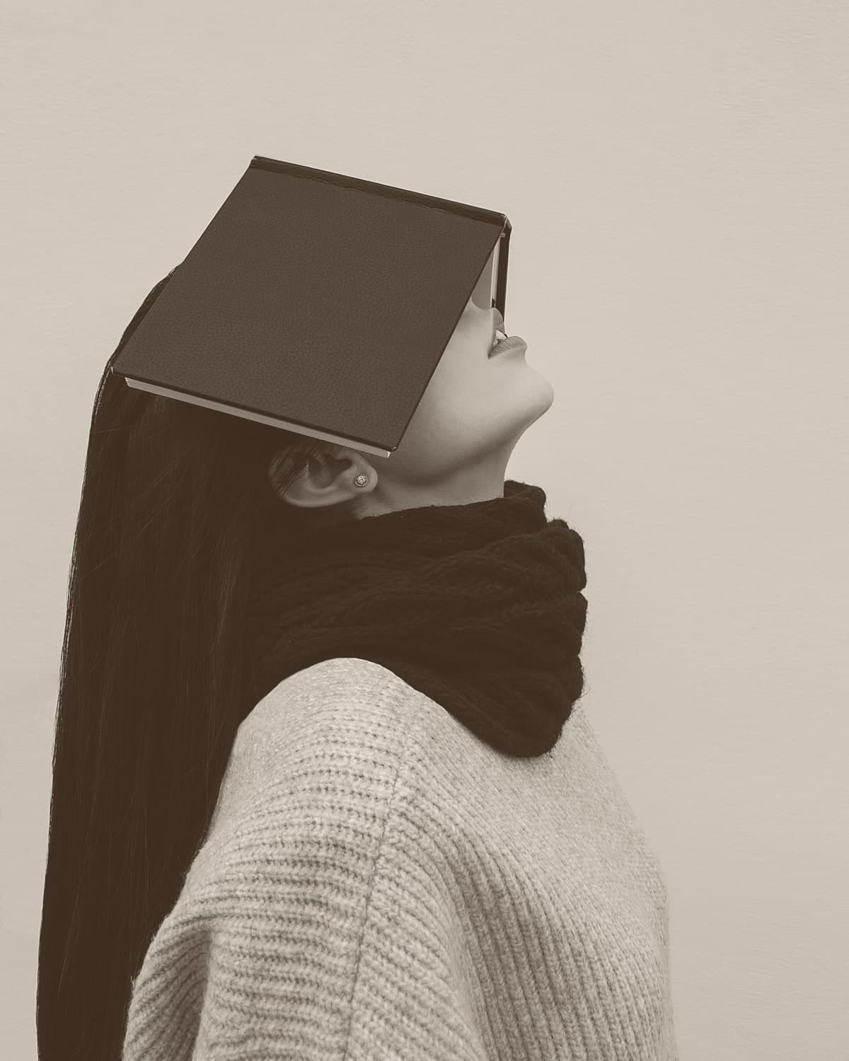 A woman in a grey knit jumper and black scarf tilts her head back with a hardcover book resting face-down over her eyes.