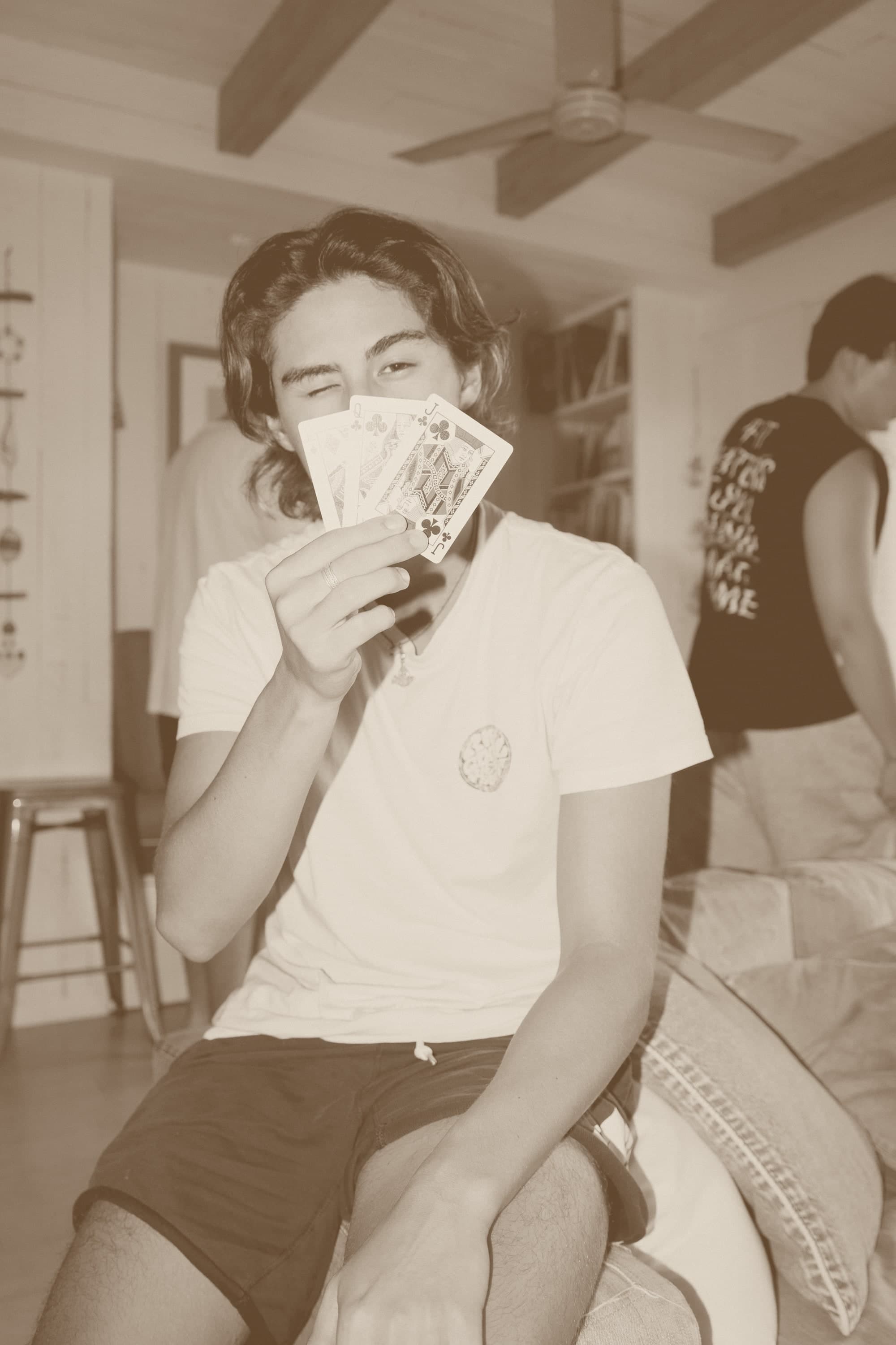 A young man holding a fan of playing cards over his face, only the eyes visible above the hand.