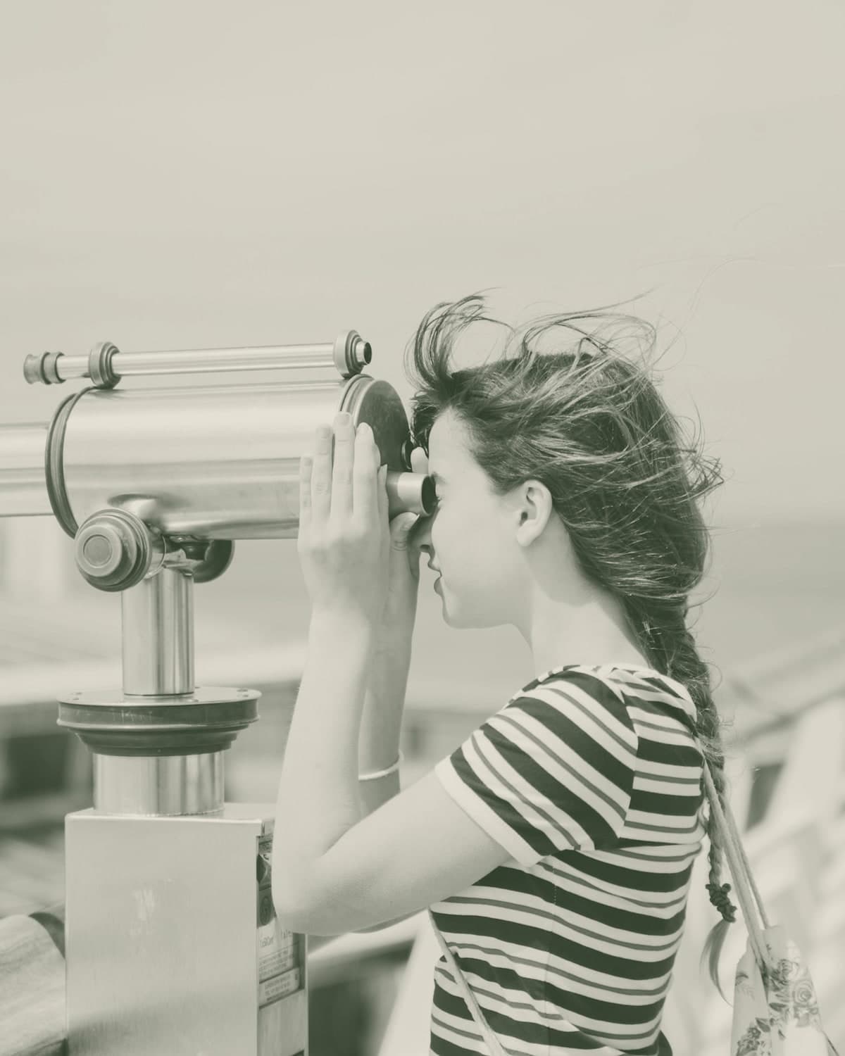 A child in a striped t-shirt leaning into a coin-operated seaside telescope, looking carefully through the eyepiece.