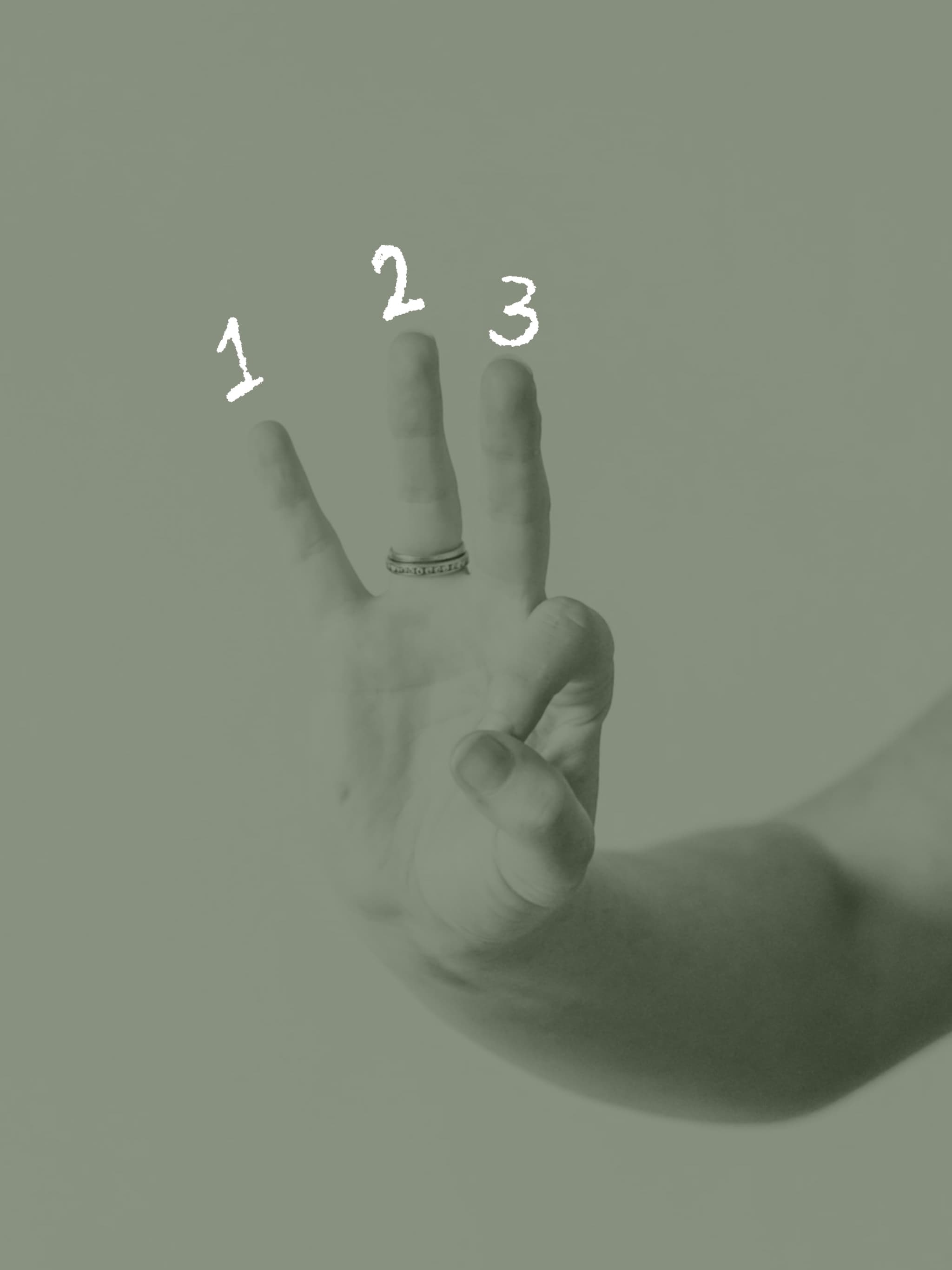 A hand raised against a soft white backdrop with three fingers held up, a slim gold ring at the base of two of them.
