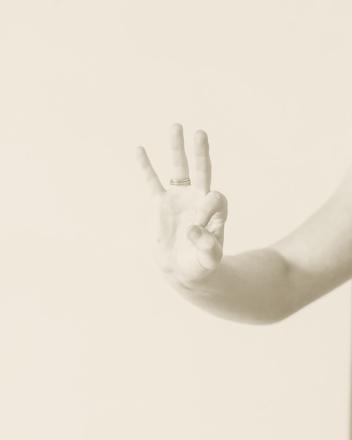 A hand raised against a soft white backdrop with three fingers held up, a slim gold ring at the base of two of them.