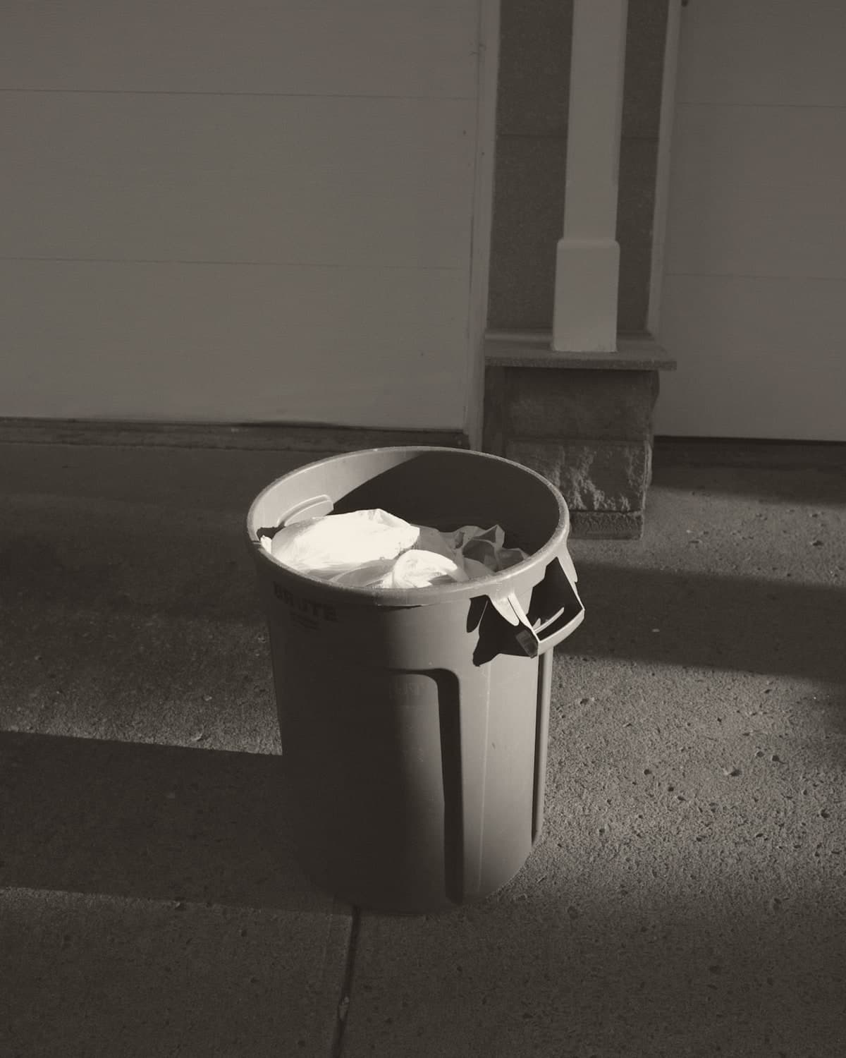 A large grey plastic bin on a sunlit pavement, a white bin bag spilling over the rim, a sharp shadow falling across the pale blue wall behind.