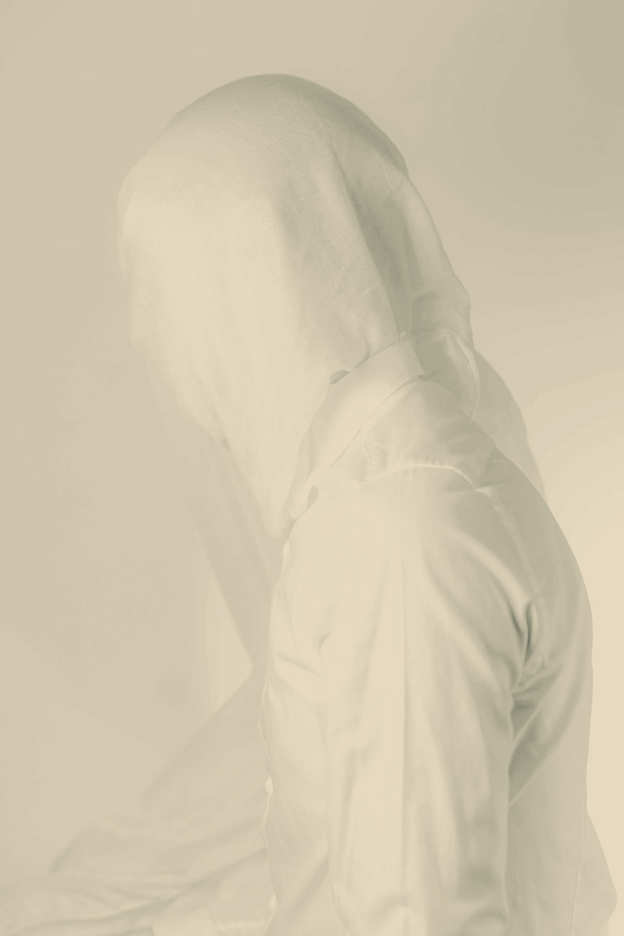 A figure in a white shirt with a hood pulled over their head, face obscured, photographed against a pale wall in soft daylight.